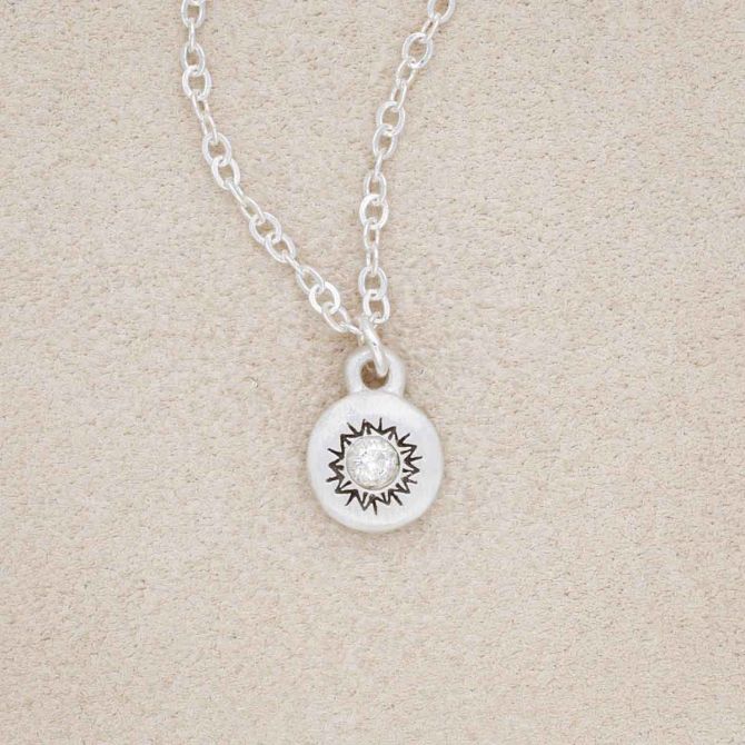 Dainty Sunburst Necklace cast in Sterling Silver and set with a 3mm cubic zirconia stone, on a beige background