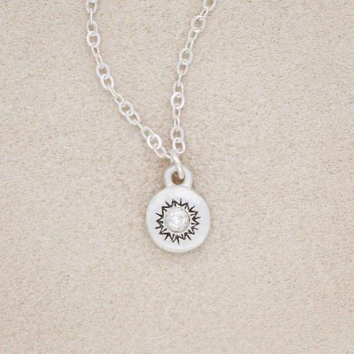 Dainty Sunburst Necklace cast in Sterling Silver and set with a 3mm cubic zirconia stone, on a beige background